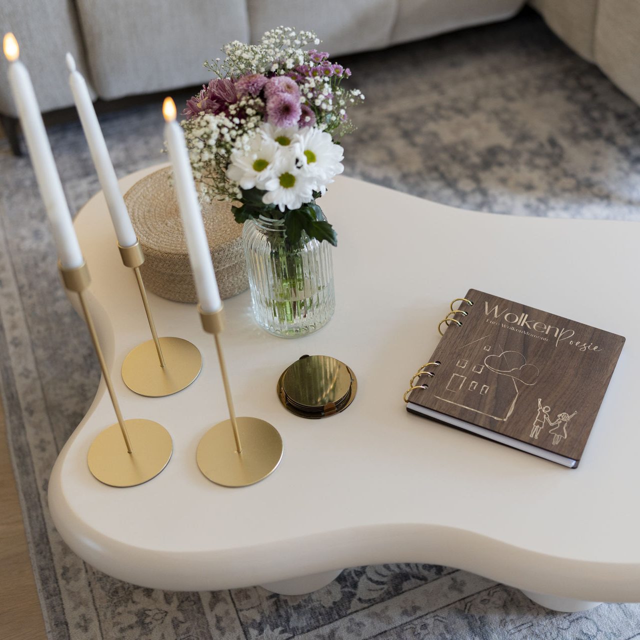 Couchtisch mit goldenen Kerzenhaltern, Blumen in einer Glasvase und einem Buch mit Holzeinband